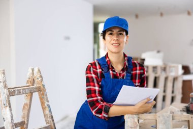 Interested smiling young female builder contractor planning renovation work at construction site, inspecting room and taking notes .. clipart