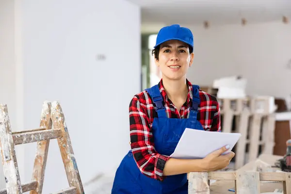 stock image Interested smiling young female builder contractor planning renovation work at construction site, inspecting room and taking notes ..