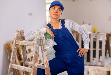 Portrait of confident young woman construction worker in blue overalls posing near wooden stepladder inside house under renovation.. clipart