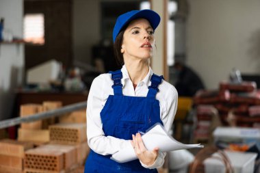 Experienced female foreman in blue workwear making task list during works in building under construction.. clipart