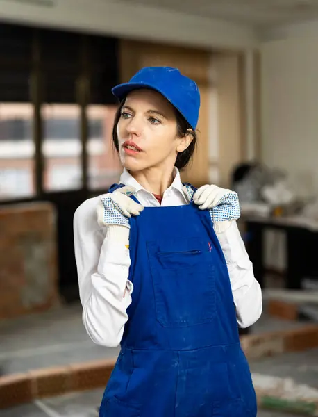 stock image Confident positive female builder wearing blue overalls and cap posing at construction site indoors..