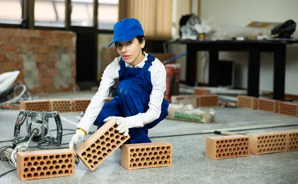 stock image Female builder in blue workwear planning erection of partition wall in building under construction, preparing red bricks for work