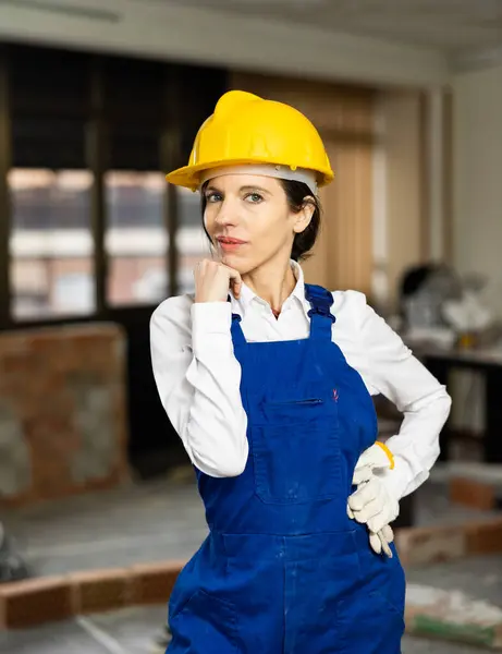 stock image Portrait of confident successful female foreman wearing blue overalls and yellow safety hard hat standing inside building under construction