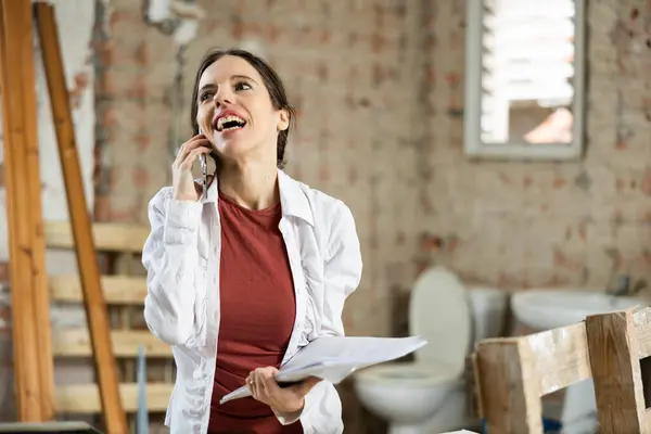 stock image Positive female designer standing in building under renovation with papers in hands, discussing workflow with customer on phone