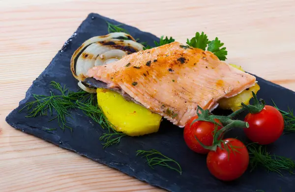 stock image Delicious baked trout fillet served on black board with potato, grilled onion rings, tomatoes and greens