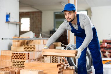 Professional builder working on a construction site indoors puts red brick construction material on top of each other clipart