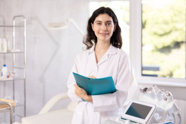 Female cosmetic doctor meticulously stands in her clinic, recording detailed notes in folder, ensuring her patients receive personalized care. clipart