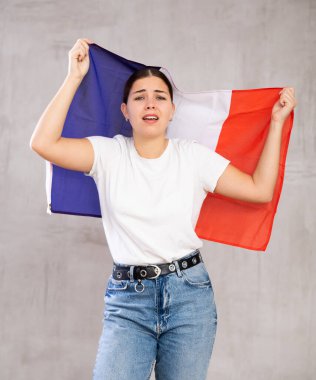 Sad young woman with France flag in hands posing sorrowfully against light unicoloured background clipart