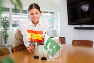 Smiling young woman putting small flag of Spain next to the flag of Pakistan placed on table in meeting room clipart