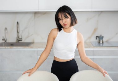 Disappointed young Asian lady standing by chairs in the kitchen with clean white cupboards clipart