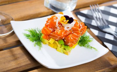 Fresh salmon tartare formed as cube with chopped avocado, onion rings, greens and pansy flowers on plate clipart