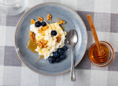 Fresh farm cottage cheese filled with large amount of liquid summer honey is served on plate, decorated with walnut and blueberry clipart