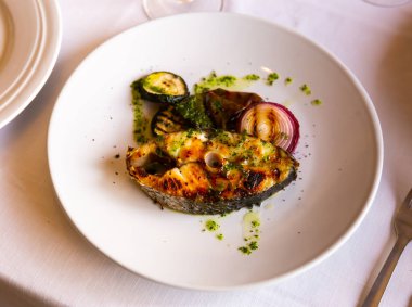 Appetizing grilled corvina steak accompanied by roasted vegetables, garnished with spicy green herb sauce, served on white plate for dinner clipart