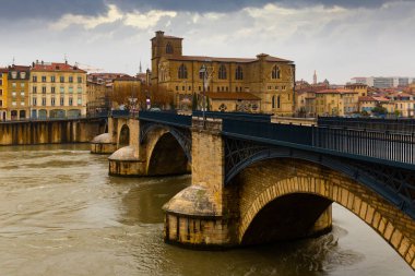 View of Romans-sur-Isere townscape on bank of Isere river with Gothique building of Collegiate church Saint Barnard and ancient arched bridge on cloudy winter day, Drome department, France.. clipart