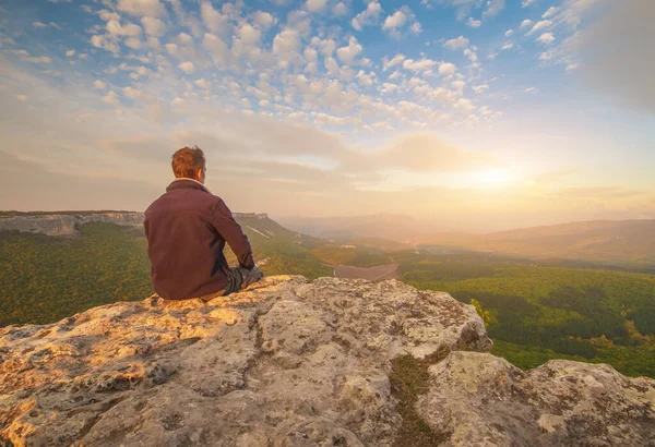 男は崖の山の端に座って太陽を見て — ストック写真
