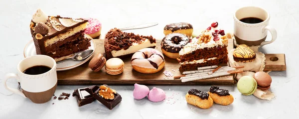 Selection of various cake pieces, macaroons, meringues, cookies and donuts on a wooden desk on a light background. Top view. Confectionery background. Panorama.