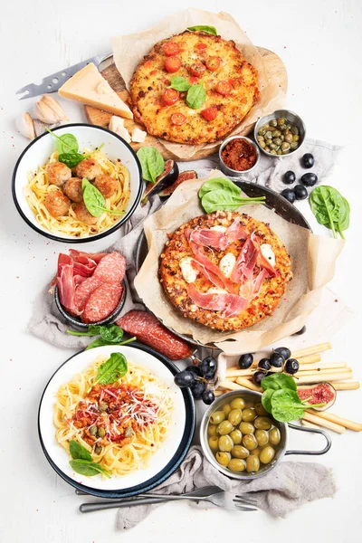 Full table of italian meals on plates  and board - Pizza, pasta, meatballs, salami, parmesan, olives, bread sticks on a white background. Top view.