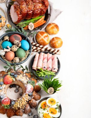 Traditional Easter dinner or  brunch with ham, colored eggs, hot cross buns, cake and vegetables. Easter meal dishes with holday decorations. Top view, copy space