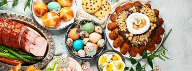 Classic Easter ham dinner. Top view table scene on a white background. Ham, eggs, hot cross buns, carrot, cake and vegetables. Panorama, banner.