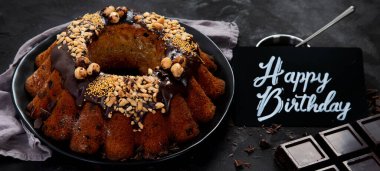 Bundt chocolate cake decorated with melted chocolate, nuts  and sprinkles on plate  on black background. Holiday concept. Top view, banner