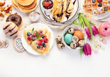 Easter sweet dessert table. Pancakes, crepes, waffles and donuts  with fresh berries, nuts and topping.   Easter traditional natural colorful eggs. Top view, copy space