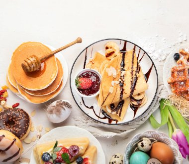 Easter sweet dessert table. Pancakes, crepes, waffles and donuts  with fresh berries, nuts and topping.   Easter traditional natural colorful eggs. Top view, copy space
