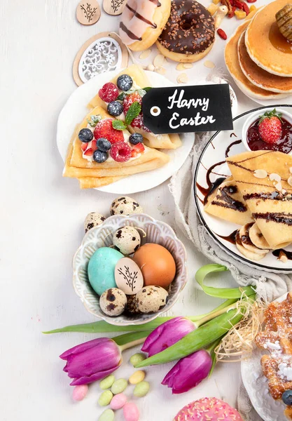 Easter sweet dessert table. Pancakes, crepes, waffles and donuts  with fresh berries, nuts and topping.   Easter traditional natural colorful eggs. Top view, copy space