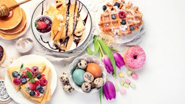 Easter sweet dessert table. Pancakes, crepes, waffles and donuts  with fresh berries, nuts and topping.   Easter traditional natural colorful eggs. Top view, copy space