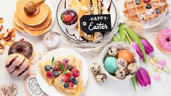 Easter sweet dessert table. Pancakes, crepes, waffles and donuts  with fresh berries, nuts and topping.   Easter traditional natural colorful eggs. Top view, flat lay