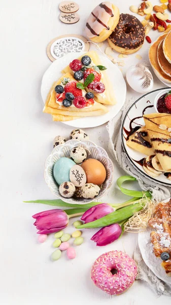 Easter sweet dessert table. Pancakes, crepes, waffles and donuts  with fresh berries, nuts and topping.   Easter traditional natural colorful eggs. Top view, copy space