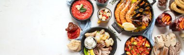 Typical spanish tapas concept. Concept include jamon, chorizo sausage, brushettas, bowl with olives, shrimp, pan with paella, cheese, sangria, churros on a white background. Top view. Panorama with copy space.