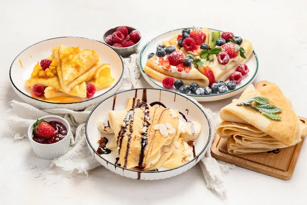 Variety of fresh crepes with chocolate, jam, fruits and berries on white background. Homemade delicious crepes. Top view.