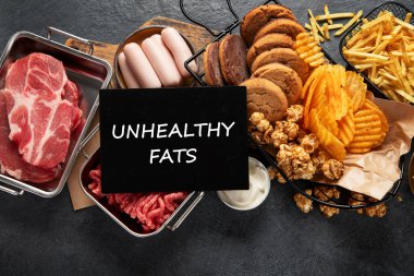 Doymuş ve trans. Sağlıksız yiyecekler. Sosis, patates, et, peynir, patlamış mısır, kurabiye, koyu arkaplanda krema. Üst görünüm.