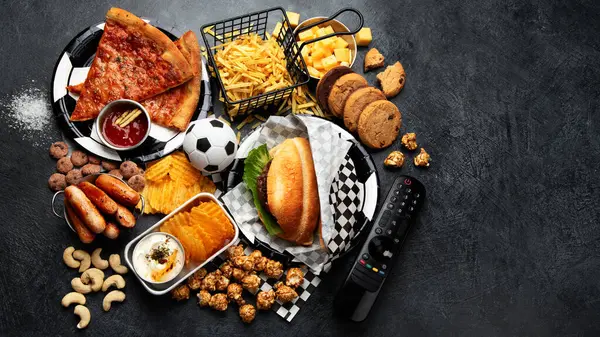 Saturated fats. Football time. TV remote control and snacks - chips, popcorn, cookies, cheese, sauce, fries, burger, nuts. Top view, copy space, banner