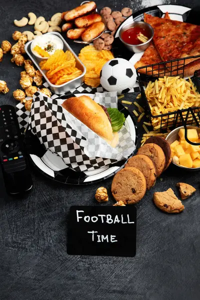 Saturated fats. Football time. TV remote control and snacks - chips, popcorn, cookies, cheese, sauce, fries, burger, nuts. Top view