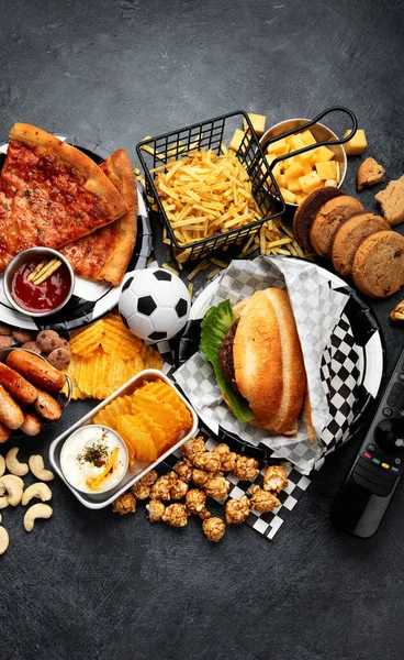 Saturated fats. Football time. TV remote control and snacks - chips, popcorn, cookies, cheese, sauce, fries, burger, nuts. Top view