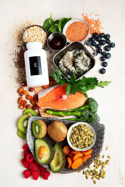 Kurutulmuş kayısı, fındık, badem, maydanoz, avokado, ceviz, kabak çekirdeği, ayçiçeği çekirdeği, ıspanak ve beyaz bir zemin üzerinde balık gibi E vitamini açısından zengin gıdalar. Üst görünüm.