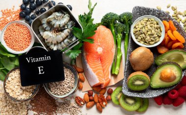 Kurutulmuş kayısı, fındık, badem, maydanoz, avokado, ceviz, kabak çekirdeği, ayçiçeği çekirdeği, ıspanak ve beyaz bir zemin üzerinde balık gibi E vitamini açısından zengin gıdalar. Üst görünüm.