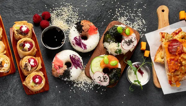 Hybrid trendy food on dark background. Sushi roll pancake, donut sushi, pizza with pasta. Top view, copy space