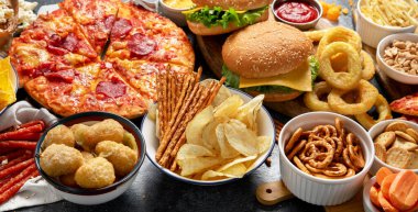 Geniş bir masa dolusu yiyecek, pizza, patates kızartması, soğan halkaları, hamburger ve atıştırmalıklar gibi karanlık arka planda. Üst görünüm.