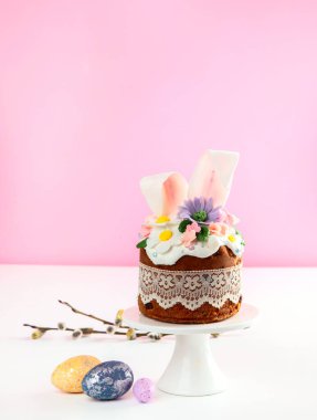 Paskalya pastası, tavşan kulaklarıyla süslenmiş tatlı ekmek ve pembe arka planda bahar çiçekleri. Üst görünüm.