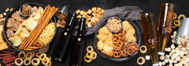 Koyu arka planda çeşitli bira ve tuzlu atıştırmalıklar. Parti yemeği konsepti. Üst manzara, panorama pankartı.