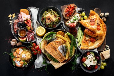 Çeşitli Akdeniz yemekleri - balık, deniz ürünleri, soslar, makarna, atıştırmalıklar, siyah arka planda pizza. Üst görünüm. 