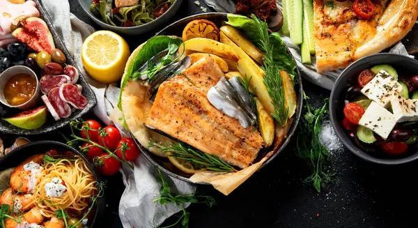 Çeşitli Akdeniz yemekleri - balık, deniz ürünleri, soslar, makarna, atıştırmalıklar, siyah arka planda pizza. Üst görünüm. 