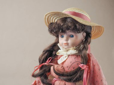 Eski model porselen bebek kız mavi gözlü, kahverengi örgülü, pembe elbiseli, beyaz önlüklü, cebinde oyuncak ve hasır şapkalı.