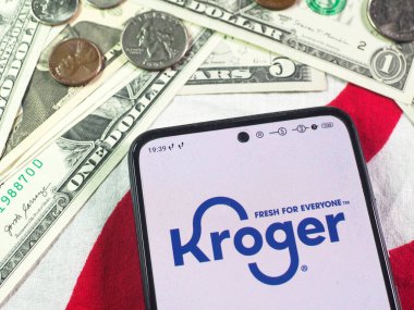 Bu fotoğraf illüstrasyonunda, Kroger Co. logosu arka planda bir akıllı telefon ve Amerikan para birimi notaları ve sikkeleri üzerinde görüntülenmektedir..