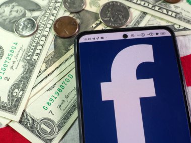 Bu resimde, Facebook logosu arka planda akıllı telefon ve Amerikan para birimi notları ve sikkeleri üzerinde gösteriliyor.