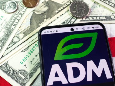 Bu fotoğraf illüstrasyonunda, Archer Daniels Midland Şirketi logosu arka planda bir akıllı telefon ve Amerikan para birimi banknotları ve sikkeleri üzerinde sergilenmektedir..