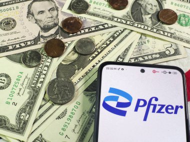 Bu fotoğraf illüstrasyonunda, Pfizer, Inc. logosu arka planda bir akıllı telefon ve Amerikan para birimi notaları ve sikkeleri üzerinde görüntülenmektedir..