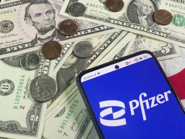 Bu fotoğraf illüstrasyonunda, Pfizer, Inc. logosu arka planda bir akıllı telefon ve Amerikan para birimi notaları ve sikkeleri üzerinde görüntülenmektedir..